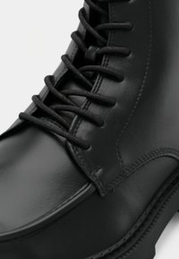 Gros plan sur une botte en cuir noir poli avec des lacets ronds noirs attachés à travers des œillets métalliques et des coutures détaillées sur le côté.