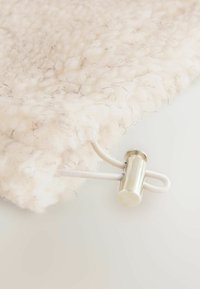 Texturiertes, cremefarbenes Material mit flauschigem Aussehen, das dünne schwarze Fäden aufweist. Beinhaltet einen silbernen Hardware-Verbinder mit weißen Kabeln.