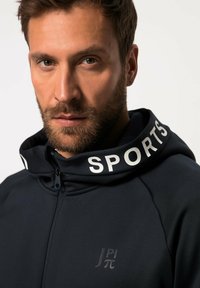 Marineblaue Sport-Hoodie mit Reißverschluss vorne, einer großen Kapuze, auf der "SPORTS" in weißem Text steht, und einem kleinen Logo auf der Brust. Glatter, moderner Stoff.