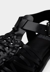 Μαύρο δερμάτινο σανδάλι με πλεγμένο σχέδιο λουριού, ρυθμιζόμενο κούμπωμα και ομαλή εσωτερική σόλα. Επισημασμένες υφές και κομψές επιφάνειες.