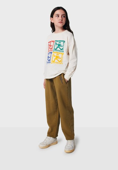 Cremesfarbene Langarmshirt mit buntem Tier-Graphic, kombiniert mit lockeren olivgrünen Hosen und weißen Sneakern mit silbernen Akzenten.