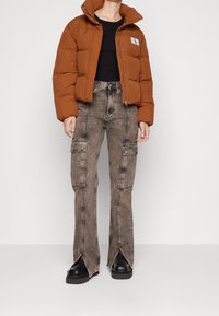 Chaqueta tipo pluma marrón con cuello alto, cierre de cremallera y parche con logo, combinada con jeans de estilo cargo descoloridos que cuentan con bolsillos laterales y dobladillos desgastados.