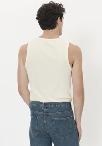 Šviesiai smėlio spalvos marškinėliai be rankovių, su apvaliu kaklu ir kreminėmis detalėmis, dėvimi su mėlynais džinsais, kurie turi standartines kišenes ir švelnią tekstūrą.