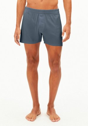 Mann trägt schlichte dunkelgraue Boxershorts und steht barfuß vor weißem Hintergrund.