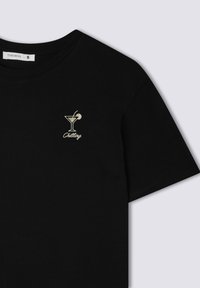 T-shirt in cotone nero con un piccolo bicchiere da cocktail ricamato in oro e la scritta "Chilling" sottostante. Design classico con collo a giro.