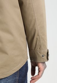 Beżowa kurtka o gładkiej fakturze, z mankietem zapinanym na zatrzask oraz białymi przeszyciami. Nosi się ją na niebieskich dżinsach.