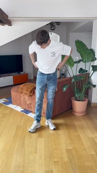 Maglietta bianca con piccolo logo, jeans strappati blu e sneakers bianche. Ambientazione interna con un divano in corduroy marrone e una pianta in vaso.
