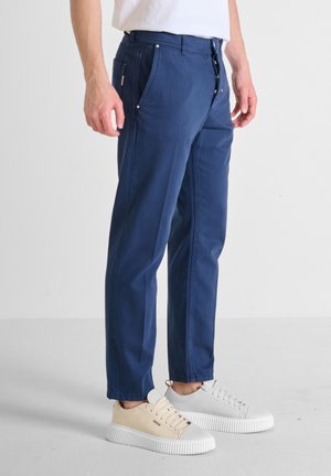 Navy blue pants made of cotton with a straight cut, featuring side pockets and metal accents, paired with light-colored sneakers.