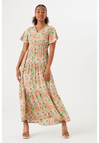 Mujer vestida con un colorido vestido maxi de cuello en V con mangas de volantes y tacones abiertos de color beige, de pie frente a un fondo blanco liso.