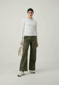 Valkoinen pitkähihainen paita, oliivinvihreät leveät housut, valkoiset tennarit, kevyesti beigen värinen laukku, neutraalia taustaa vasten.