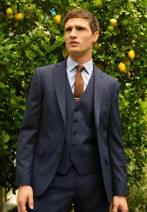 Traje de tres piezas en azul marino con un acabado texturizado, que presenta una solapa con muesca, cinco botones y una corbata con lunares, sobre un fondo verde.
