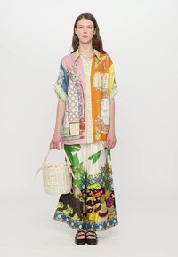Lysfarvet oversized skjorte med blomster- og vinduesmotiver, parret med mønstrede, lette bukser. Modellen holder en vævet taske.