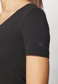 Crna kratka majica s kratkim rukavima i V izrezom, izrađena od mekog materijala, s diskretnim logotipom na lijevom rukavu. Glatka tekstura, uski kroj.