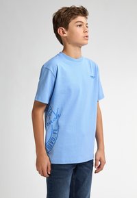 Camiseta de algodón de color azul claro con mangas cortas. Presenta un gráfico circular impreso en azul oscuro en el lado izquierdo y un pequeño logo en el pecho.