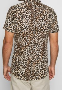 Mand iført skjorte med korte ærmer i beige, sort og brun leopardmønster, kombineret med lyse bukser, vist fra ryggen.