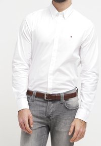 Witte blouse met lange mouwen en een klein logo. Gecombineerd met grijze jeans en een bruine leren riem.