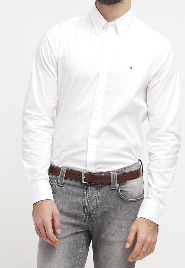 Camicia bianca con bottoni e maniche lunghe, con un piccolo logo. Abbinata a jeans grigi e una cintura di pelle marrone.