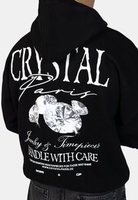 Czarna bluza z kapturem z białym napisem "CRYSTAL Paryż" oraz dużymi grafikami diamentów, dodatkowy tekst o biżuterii i zegarkach z tyłu.