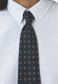 Silkeslips i mörkblått med diamantmönster som har blommiga inslag i brunt, vitt och gult, tillsammans med en vit, knappad skjorta.