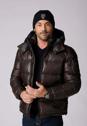 Homme portant un bonnet en maille noir, une doudoune marron foncé sur un pull noir, ajuste ses manches, sur un fond gris clair uni.