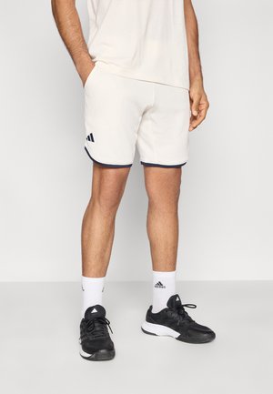 Herren-Basketballshorts in Beige mit marineblauem Rand und drei gestreiften Akzenten, kombiniert mit schwarzen Sportschuhen und weißen Socken. Glatte Textur.