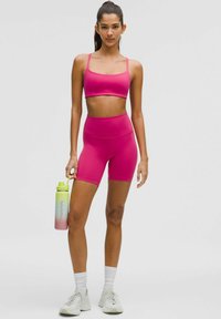 Set sportiv de culoare roz aprins, format dintr-un top scurt și pantaloni scurți cu talie înaltă. Modelul ține în mână o sticlă de apă cu gradient și capac galben.