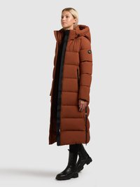 Hnědý nadýchaný kabát s kapucí, černými zipy, dlouhým střihem a bočními kapsami. Nošený s černými kotníkovými botami.