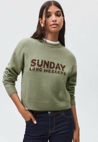 Zeleni pleteni džemper s okruglim izrezom; sadrži natpis "NEDJELJNI DOLGI VIKEND" u kontrastnom smeškastom tekstu. Mekana tekstura, rebrasti manžeti i rub.