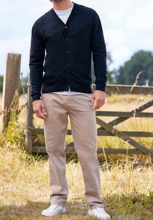 Cardigan bleu marine avec col en V et cinq boutons, porté avec un pantalon beige clair et des baskets blanches. Herbe et clôture en bois en arrière-plan.