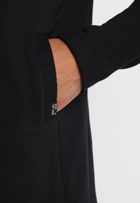 Zwarte lange mouw kleding met een zijzak en een label dat leest "DRAAG DE CODE." De stof lijkt zacht en glad met een getailleerd ontwerp.