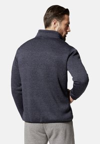 Mann, der von hinten zu sehen ist, trägt eine dunkelblaue Fleecejacke mit hohem Kragen und hellgraue Hose, steht vor einem einfarbigen Hintergrund.