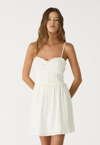 Vit sommarklänning med spagettiband, texturerad tyg, knappdetaljer och volangtopp. Har florala mönster präglade genom hela klänningen.