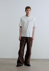 Λευκό κοντομάνικο τ-shirt με διακριτικό λογότυπο στο στήθος, συνδυασμένο με φαρδιά καφέ παντελόνια και μαύρα παπούτσια με χαμηλό τακούνι. Απλό φόντο.