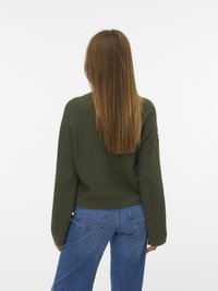 Persoon met lang, steil bruin haar, gekleed in een olijfgroene gebreide trui en spijkerbroek, met de rug naar de camera gekeerd tegen een witte achtergrond.