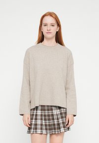 Μπεζ πολύ φαρδιά πουλόβερ με ριμπ στο λαιμό, συνδυασμένο με φούστα καρό σε καφέ και λευκούς τόνους, με πιέτες.