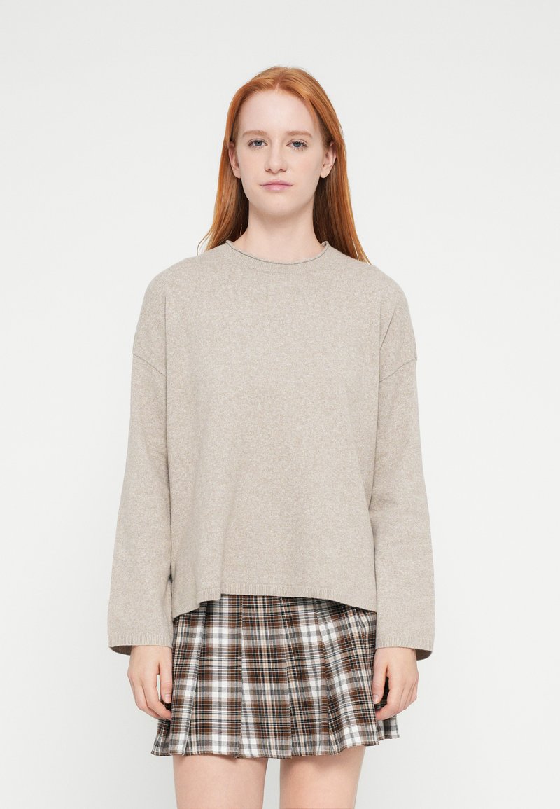 Μπεζ πολύ φαρδιά πουλόβερ με ριμπ στο λαιμό, συνδυασμένο με φούστα καρό σε καφέ και λευκούς τόνους, με πιέτες.