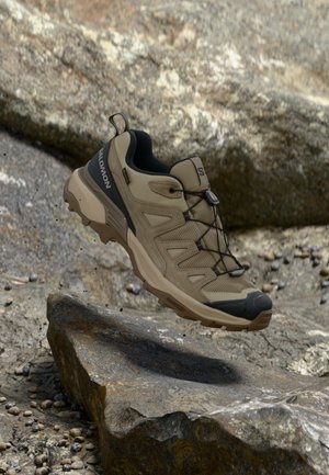 Lette beige vandresko med et mesh-overmateriale, sorte accenter, elastiske snørebånd og en robust sål, designet til udendørs brug.