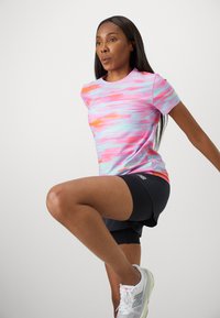 Short-sleeve athletic shirt in pink, orange, and blue pattern; paired with black shorts. The fabric appears smooth and stretchy.
