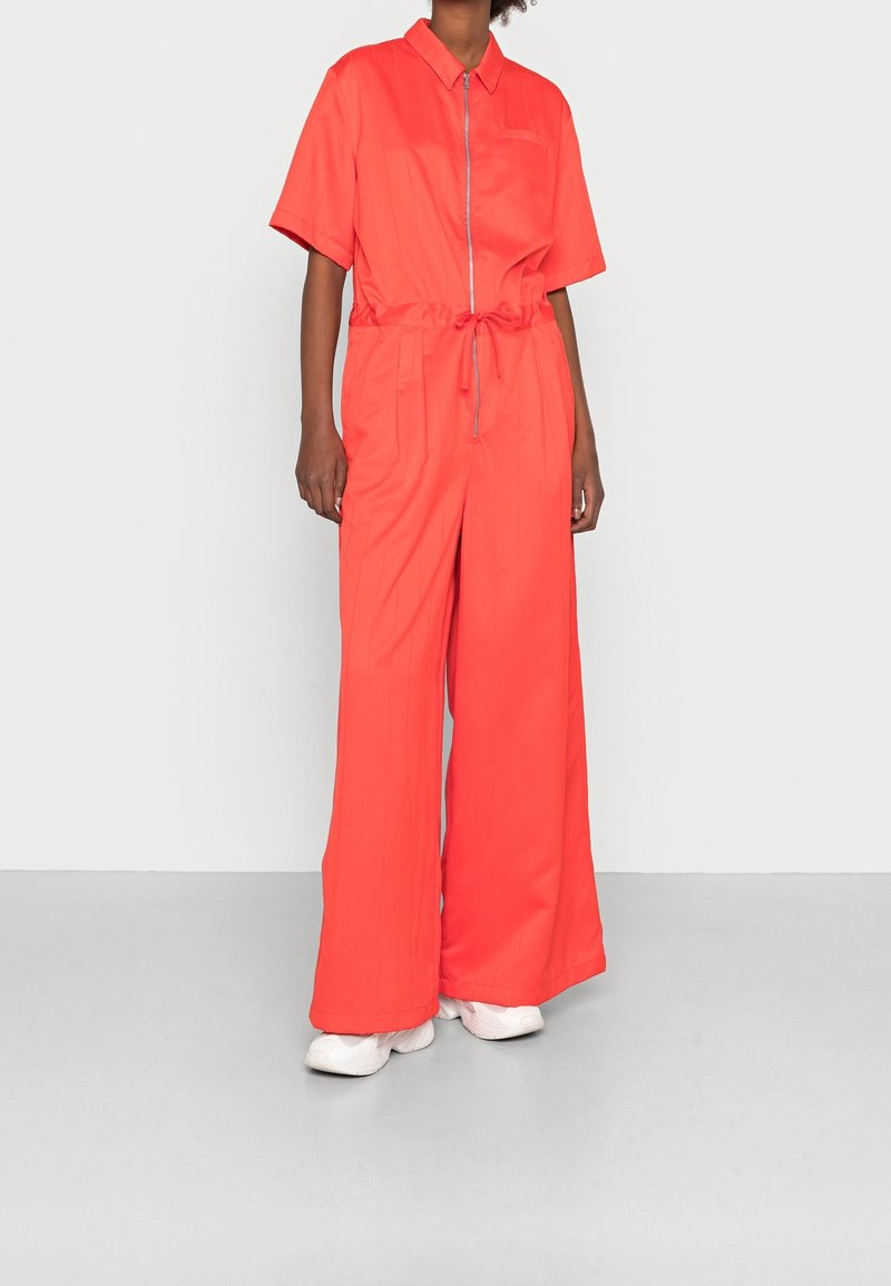 Personne portant une combinaison orange vif à manches courtes avec une fermeture éclair à l'avant et une taille à cordon, associée à des baskets blanches.