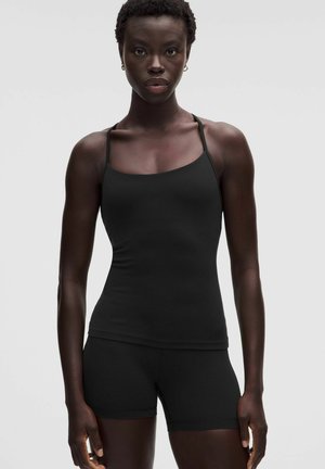 Kvinde iført sort tætsiddende tanktop og matchende shorts, stående mod en ensfarvet lys grå baggrund.