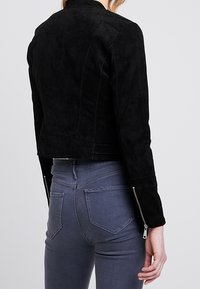 Svart mockajacka med kort längd och långa ärmar, dragkedjesmycken vid handlederna och sömnadsdetaljer. Bärs med åtsittande grå jeans.