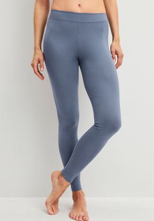 Close-up van een persoon die strakke blauwe leggings draagt, blootsvoets staat met één been licht gekruist voor het andere tegen een effen achtergrond.