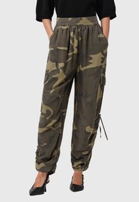 Donna che indossa pantaloni cargo mimetici verde oliva con tasche laterali e scarpe con tacco nere, con le mani nelle tasche, su uno sfondo semplice.
