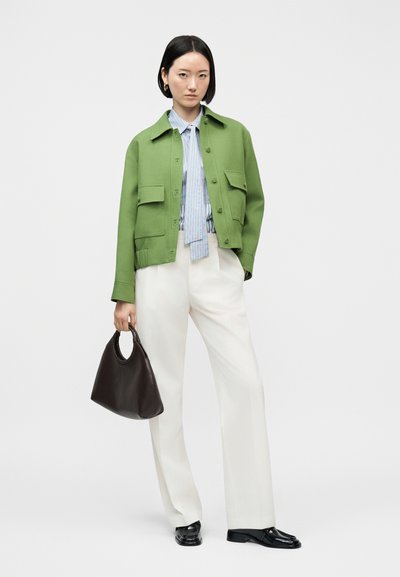 Femme portant une veste verte, une chemise rayée bleue, un pantalon blanc, des chaussures noires, et tenant un sac à main noir, debout devant un fond uni.