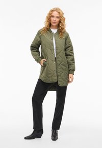 Olivgrön vadderad jacka med tryckknappar, fickor på sidorna och ribbstickade muddar, kombinerad med svarta byxor med raka ben och ankelboots.