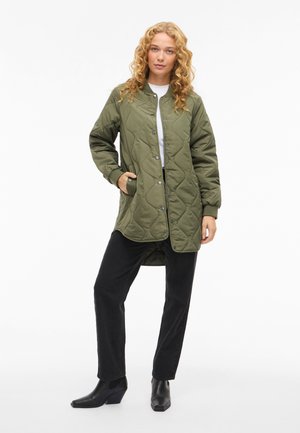 Chaqueta acolchada verde oliva con botones a presión, bolsillos laterales y puños de canalé, combinada con pantalones negros de pierna recta y botines.