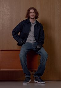 Svart jakke, grå polo-skjorte, blå jeans, svarte hansker og svarte joggesko. Satt på et trestykke med en glatt finish.