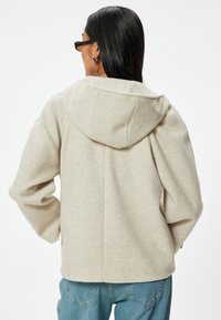 Beige huvtröja i mjukt, texturerat material, med en avslappnad passform och enkla sömmar bak, kombinerad med blå denimjeans.