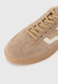 Baskets en daim beige avec des lacets marron et une semelle en gomme texturée. Présente des accents latéraux blancs et des détails de surpiqûres sur la tige.