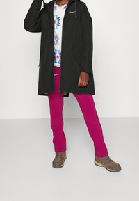 Zwarte waterdichte lange jas met capuchon, gedragen over een patroonhemd. Felroze broek en bruine wandelschoenen maken de outfit compleet.